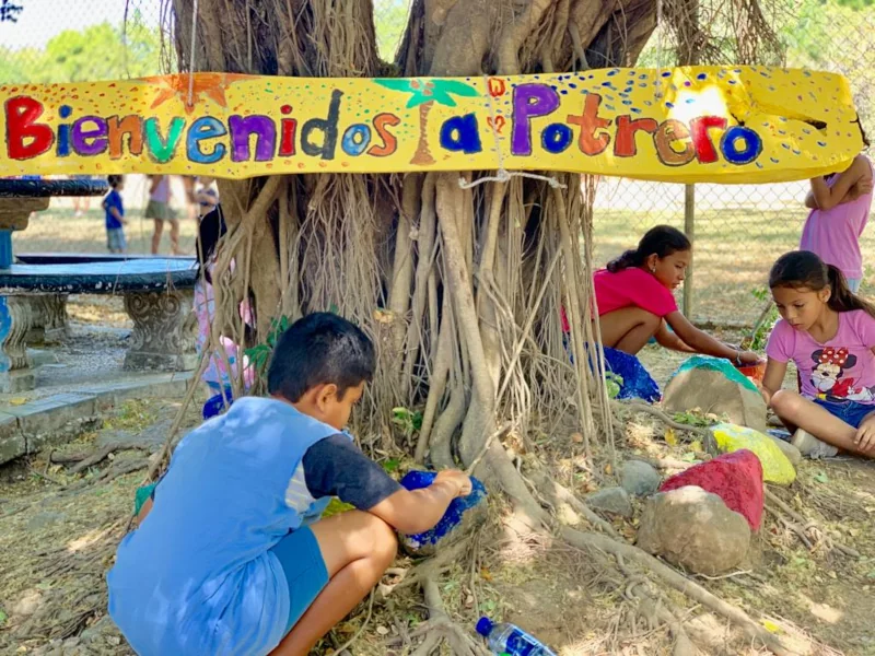 Local children painting rocks beneath a hand-painted "Bienvenidos a Potrero" sign in the community