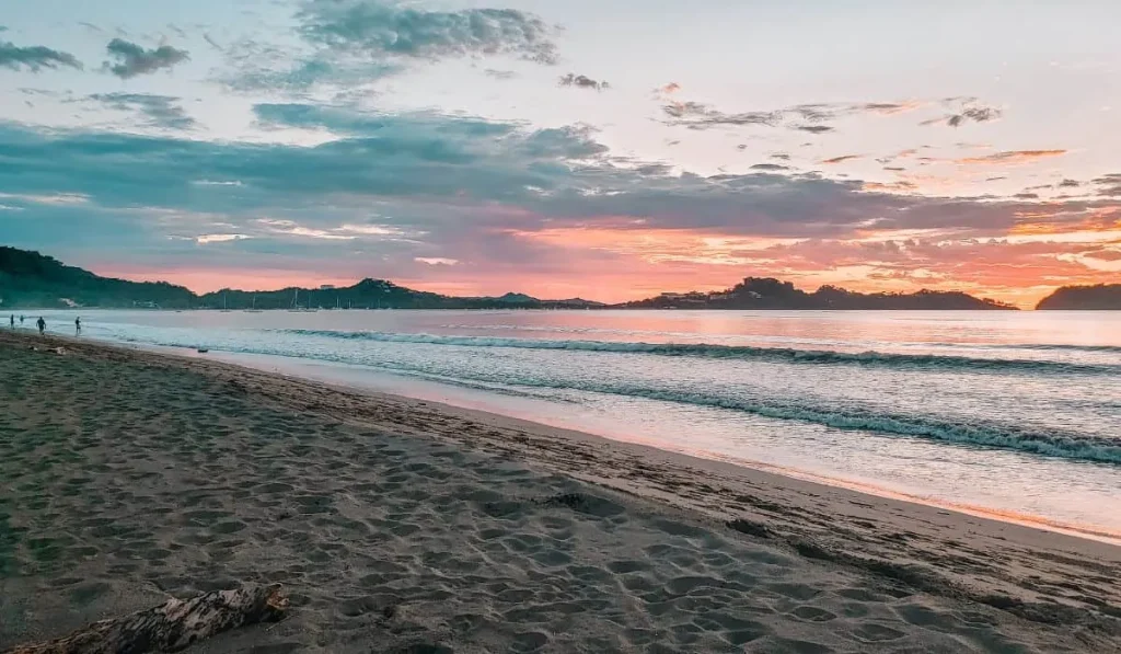 A pastel sunset over the calm bay at Playa Potrero, with empty sand, gentle surf, and the headland in the distance