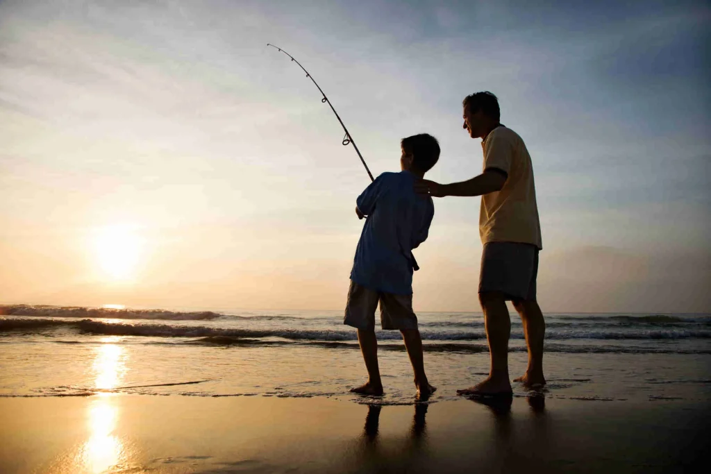 A father and son surf fishing together at sunset on a Playa Potrero beach, silhouetted against the golden sky