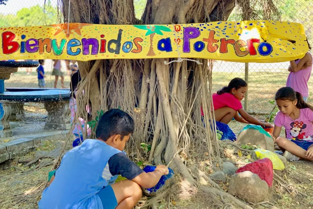 Local children painting rocks beneath a hand-painted "Bienvenidos a Potrero" sign at a community event in Playa Potrero
