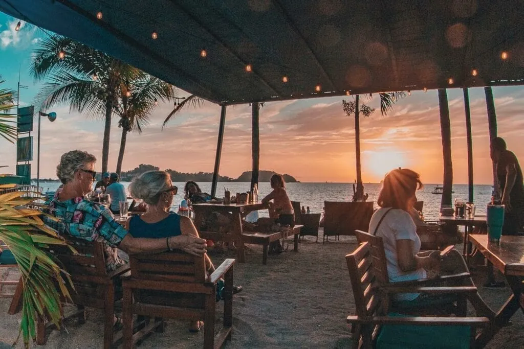 Diners at beachfront wooden tables watching the sunset over Playa Potrero, string lights and palms framing the bay