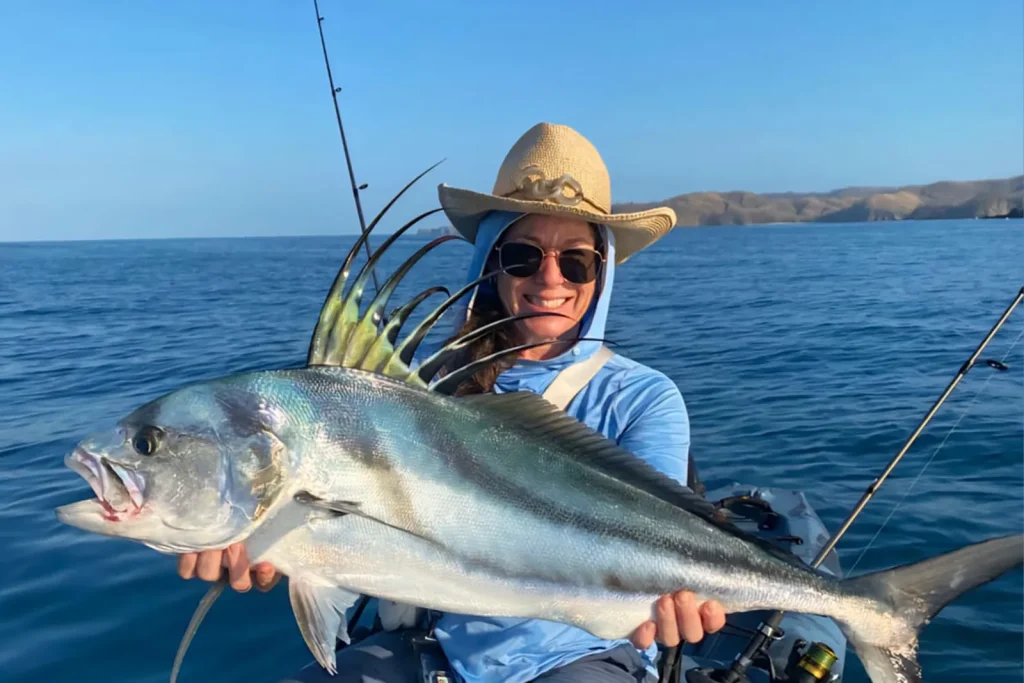 Angler in a sun hat holding a large roosterfish with its comb raised, seated in a fishing kayak with the Guanacaste coastline in the background